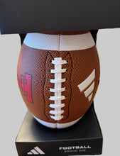 Load image into Gallery viewer, An Adidas football displayed on a black stand with its retail box. The ball is brown with white laces and white stripes, featuring a pink and white 'H' logo on the left and a white three-stripe logo on the right.
